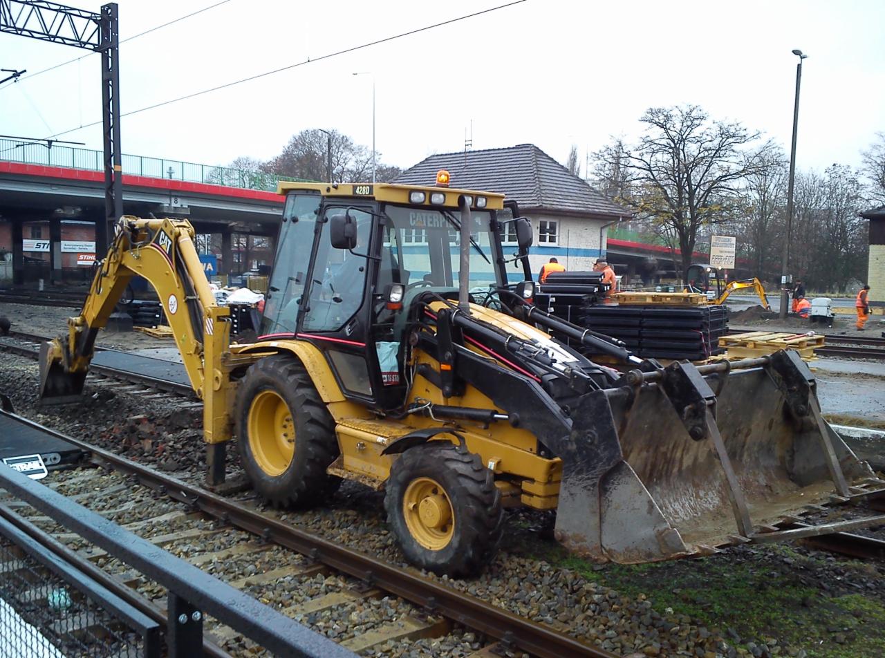 Żółta koparko-ładowarka Caterpillar na torach kolejowych, w tle budynek dworca i pracownicy w pomarańczowych kombinezonach. Widoczne elementy infrastruktury kolejowej.