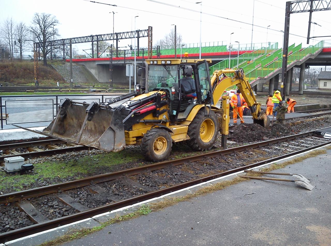 Żółta koparko-ładowarka CAT przy torach kolejowych, robotnicy w pomarańczowych kombinezonach wykonują prace ziemne. W tle widoczny wiadukt dla pieszych.