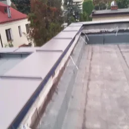 Płaski dach pokryty papą termozgrzewalną z nowymi obróbkami blacharskimi na krawędziach, widoczne budynki w tle.