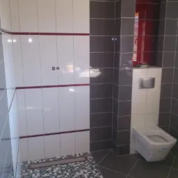 Nowoczesna łazienka z białymi i szarymi płytkami, akcentem czerwieni oraz mozaiką pod prysznicem. Podwieszana toaleta z zabudowaną spłuczką.