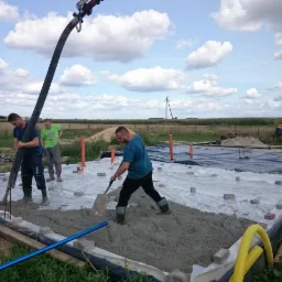 Wylewka betonowa fundamentów domu jednorodzinnego: pracownicy rozprowadzają beton szpadlami, widoczna pompa do betonu, folia izolacyjna, obrzeża z kostki, rury kanalizacyjne, teren budowy w tle.