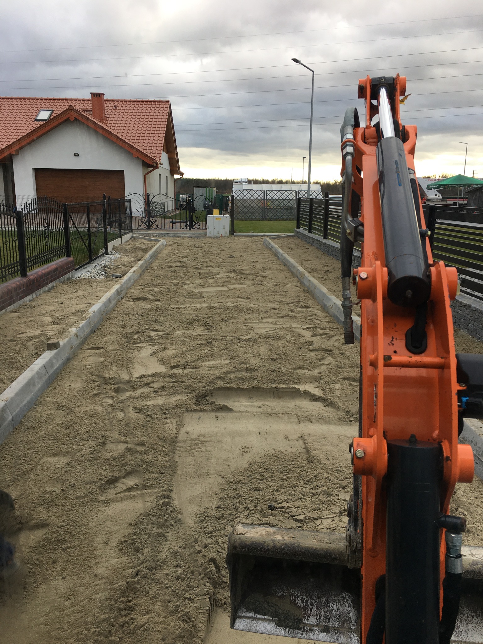 Przygotowanie podłoża pod drogę dojazdową, widoczne obrzeża betonowe i piasek wyrównywany małą koparką, w tle dom jednorodzinny.