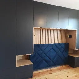 Czarna zabudowa szaf z drewnianymi wstawkami i tapicerowanym panelem ściennym w romby, uzupełniona o lampki nocne.