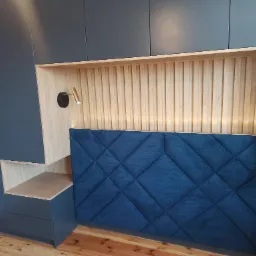 Szara zabudowa meblowa z szafami i półkami, drewniane lamele na ścianie za wezgłowiem łóżka, tapicerowany na niebiesko zagłówek z geometrycznym wzorem, oświetlenie punktowe.