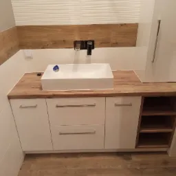 Biała, prostokątna umywalka nablatowa z baterią ścienną, zamontowana na drewnianym blacie z białymi szafkami i otwartymi półkami. Tło stanowią białe i drewnopodobne płytki ścienne.