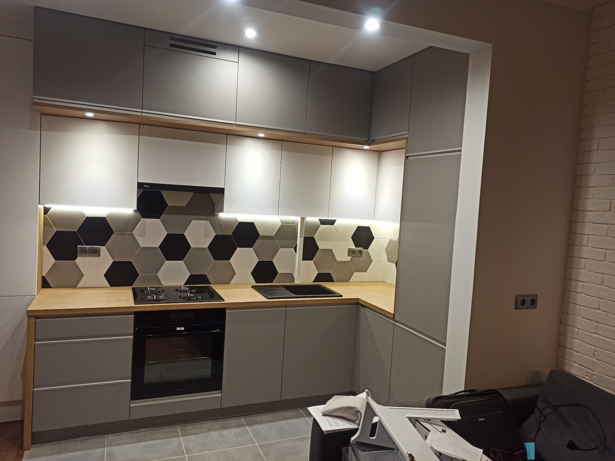 Szara kuchnia na wymiar z białymi szafkami górnymi, drewnianym blatem i heksagonalnymi płytkami na ścianie, oświetlona punktowymi lampami sufitowymi i ledami pod szafkami.