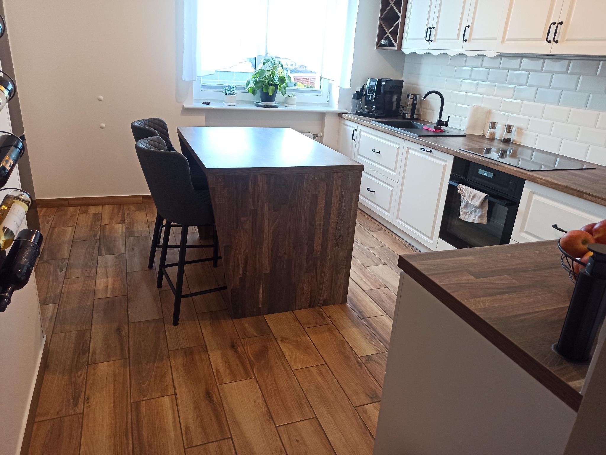 Jasna kuchnia z drewnianym blatem, wyspą i podłogą w odcieniach brązu, widok na okno z roślinami doniczkowymi.