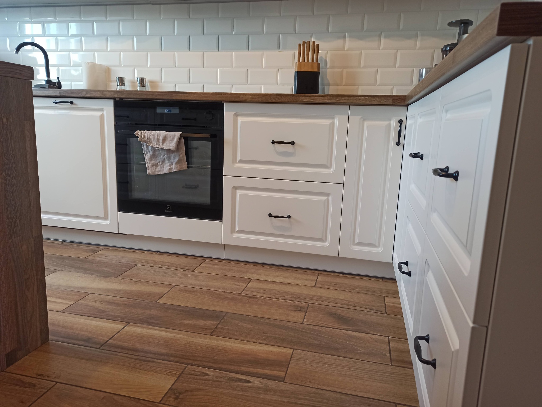 Białe szafki kuchenne z ciemnymi uchwytami, drewniany blat, piekarnik z ręcznikiem w środku, płytki imitujące drewno na podłodze i białe płytki na ścianie.
