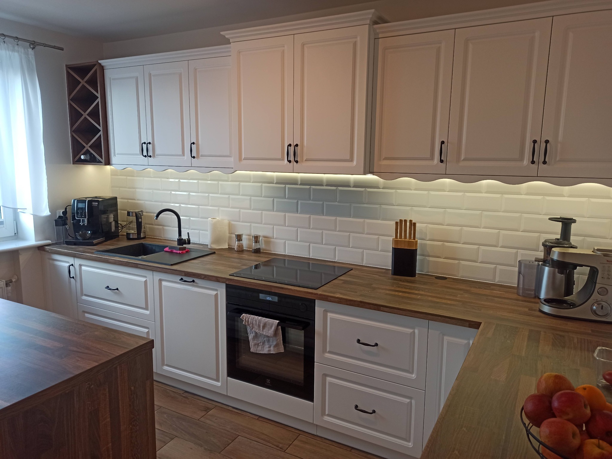 Jasna kuchnia z drewnianym blatem, białymi szafkami z frezowanymi frontami i czarnymi uchwytami, oświetlona listwą LED pod szafkami wiszącymi, z widocznym ekspresem do kawy, zlewem, płytą...