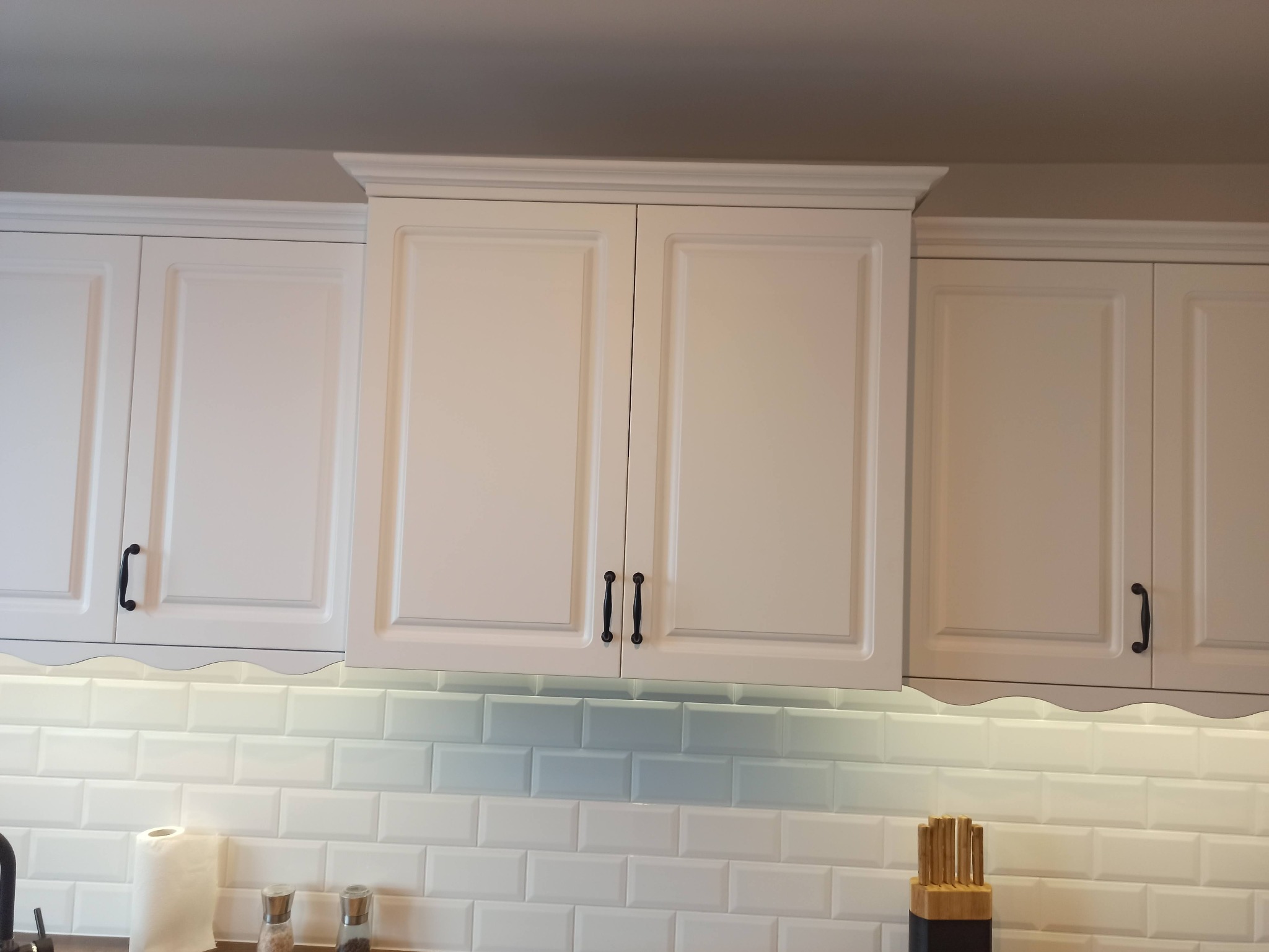 Białe szafki kuchenne z frezowanymi frontami i czarnymi uchwytami, oświetlenie podszafkowe podkreśla białe, prostokątne płytki na ścianie.