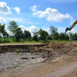 Żółta koparka CAT M 315C przy wykopie pod fundamenty domu na tle błękitnego nieba z chmurami i zieleni drzew. Widoczne ślady opon na ziemi i drewniane szalunki.