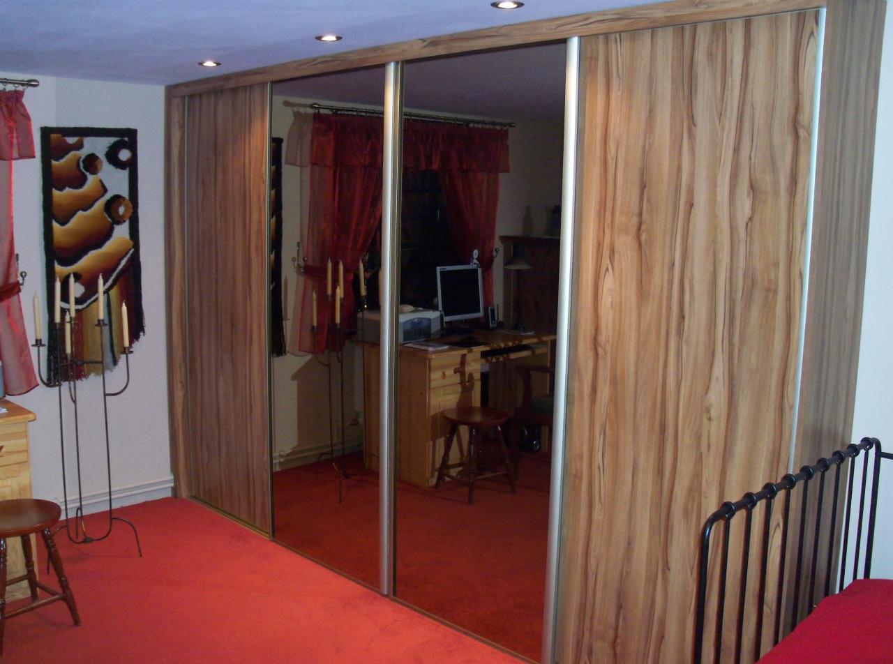 Trzydrzwiowa szafa wnękowa z lustrami, wykończona drewnopodobną okleiną. W tle widoczny pokój z czerwonym dywanem i biurkiem.