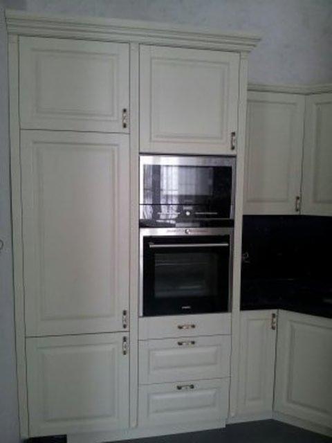 Jasna zabudowa kuchenna z frezowanymi frontami, piekarnikiem i mikrofalówką w zabudowie. Klasyczny styl wykończenia, widoczne detale frezowania na frontach szafek.