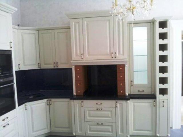 Jasna kuchnia na wymiar z ciemnym blatem i oświetleniem. Górne szafki z dekoracyjnymi uchwytami, dolne z szufladami. Nad blatem rząd małych, brązowych szufladek.