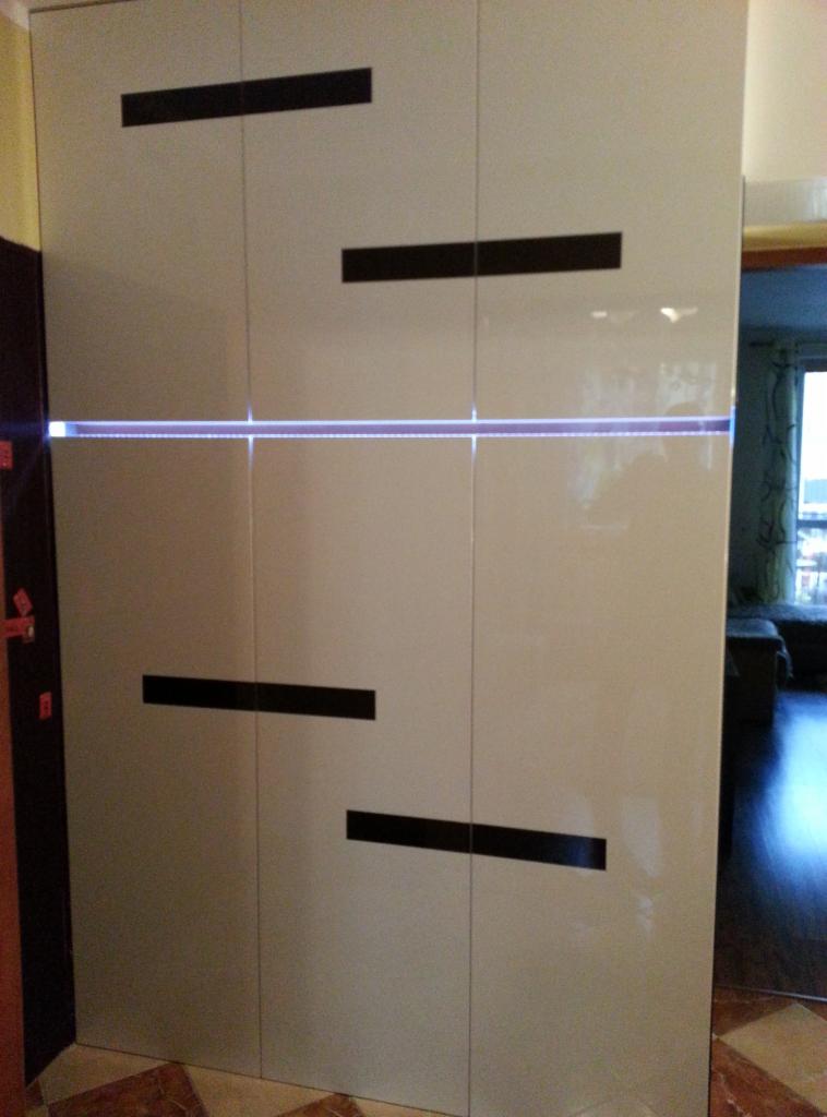 Biała szafa wnękowa z podświetleniem LED i poziomymi, czarnymi uchwytami, widoczna z perspektywy korytarza, z fragmentem podłogi z płytek.