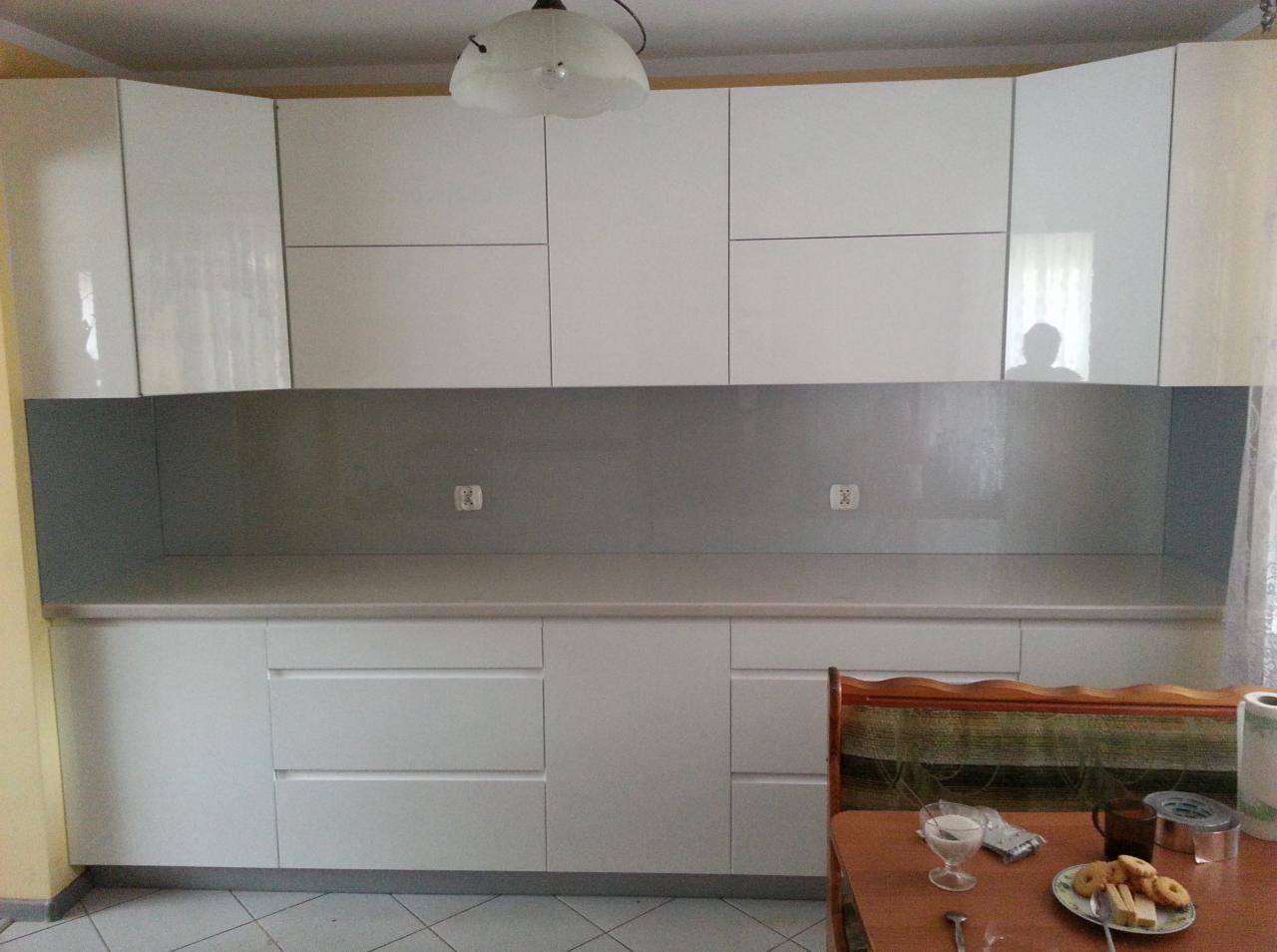 Nowoczesna, biała kuchnia z szarym panelem ściennym, widok na blat i szafki. Na stole zastawa i przekąski, wnętrze oświetlone lampą sufitową.