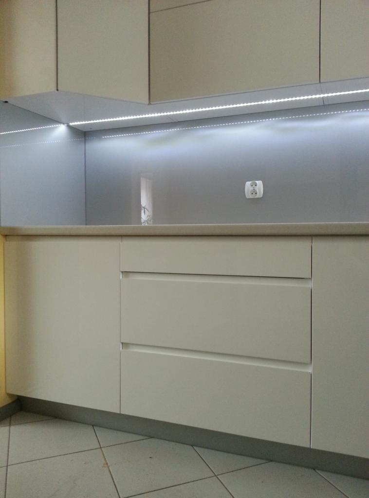 Jasna zabudowa kuchenna z szufladami, podświetleniem LED i gniazdkiem elektrycznym na szarej ściance. Minimalistyczny design, neutralna kolorystyka.