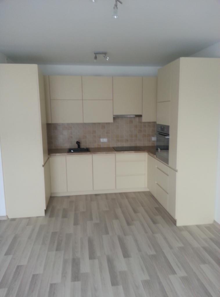 Jasna, minimalistyczna kuchnia z beżowymi frontami, widoczna w całości, z drewnianą podłogą. Uwagę zwraca brak uchwytów i gładkie powierzchnie szafek.