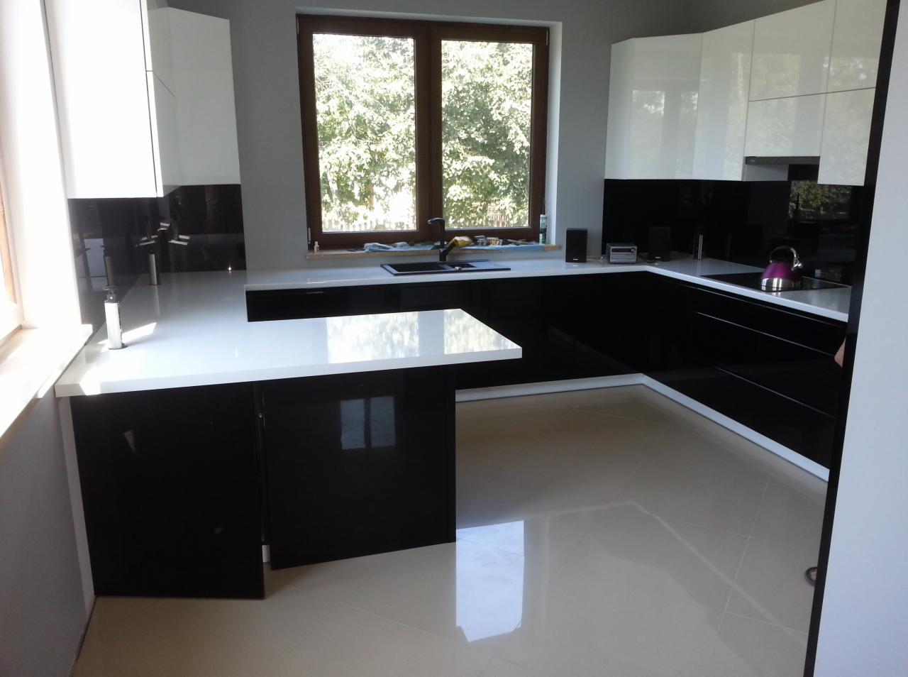 Nowoczesna kuchnia w czerni i bieli, z błyszczącymi frontami i białym blatem, na tle okna z widokiem na zieleń. Minimalistyczny design, gładkie powierzchnie, jasna podłoga.