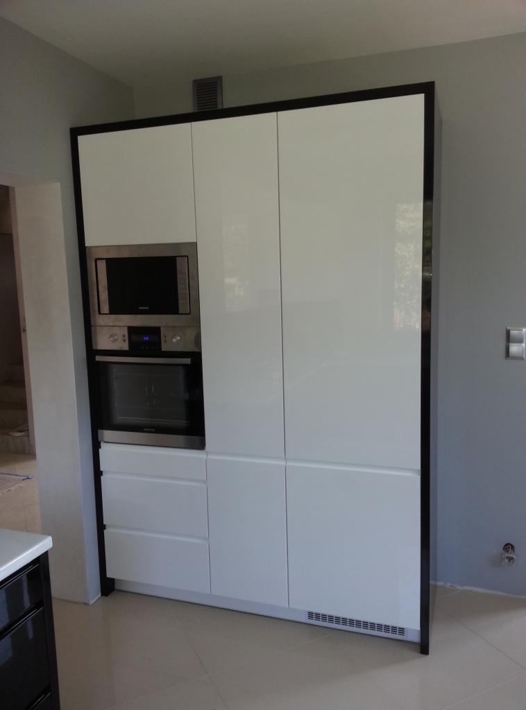 Biała zabudowa kuchenna z czarnymi obramowaniami, wbudowany piekarnik i mikrofalówka, minimalistyczny design, gładkie fronty, jasna podłoga.