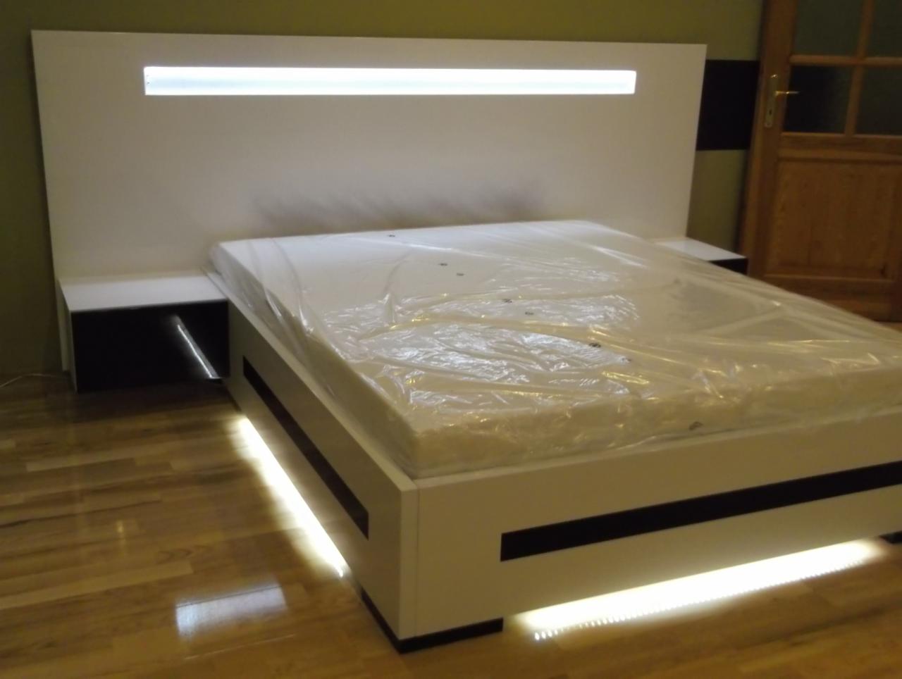 Nowoczesne łóżko z podświetleniem LED: biała rama z czarnymi akcentami, podświetlany zagłówek i szafki nocne, materac owinięty folią ochronną, drewniana podłoga.