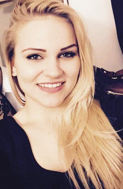 Portret uśmiechniętej blondynki z długimi włosami, ubrana w czarną bluzkę, z delikatnym makijażem, na rozmytym tle.