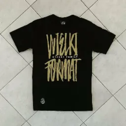 Czarna koszulka z złotym nadrukiem 'WIELKI STREET FAMILY FORMAT' leżąca na podłodze z jasnych płytek.