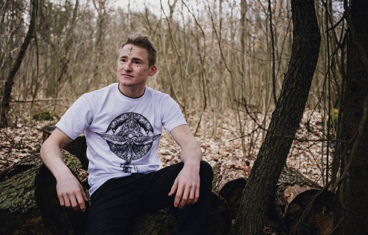 Młody mężczyzna w białej koszulce z graficznym nadrukiem, siedzący na pniu drzewa w lesie, z ozdobnym makijażem na czole.
