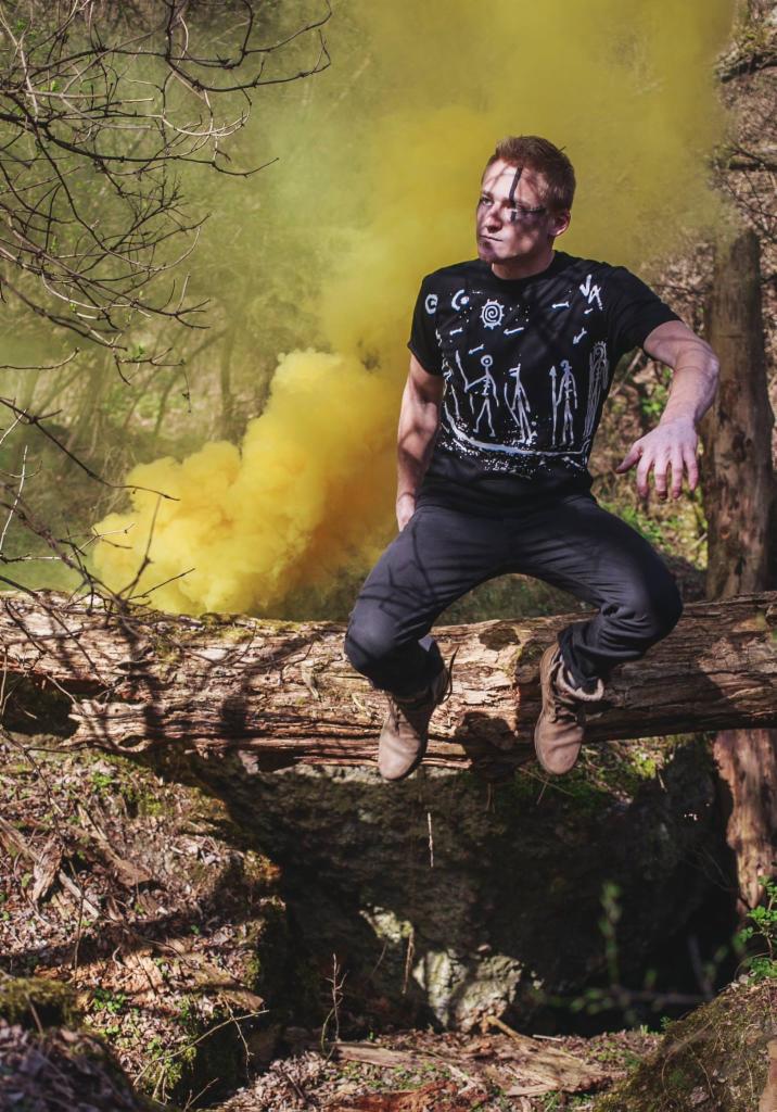 Mężczyzna w czarnej koszulce z białym nadrukiem, siedzący na powalonym pniu drzewa na tle żółtej smugi dymu w lesie.