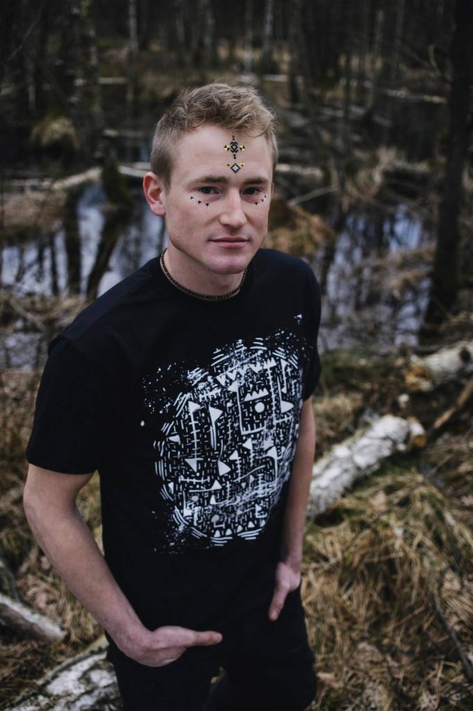 Młody mężczyzna w czarnym T-shircie z białym geometrycznym nadrukiem, stojący na tle leśnego krajobrazu z elementami wody, z ozdobami na twarzy.