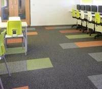 Wykładzina dywanowa w biurze, ułożona z kwadratowych płytek w kolorach szarości, zieleni i pomarańczu, z widocznymi krzesłami w kolorze limonkowym i stołami z jasnego drewna.