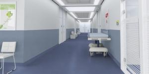 Korytarz szpitalny z niebieską wykładziną podłogową, białymi ścianami i meblami. Widoczny wózek medyczny i krzesło.