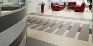 Wnętrze biurowe z jasną wykładziną w geometryczny wzór szarych i czerwonych prostokątów, czerwone sofy w tle, recepcja z lewej strony.