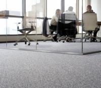 Jasnoszara wykładzina dywanowa w biurze, widoczne szklane ścianki działowe i sylwetki osób przy stole konferencyjnym za nimi.