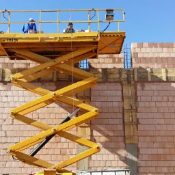 Dwóch pracowników na podnośniku nożycowym H15 SX, pracujących przy wznoszeniu muru z cegły ceramicznej, z widocznym szalunkiem i fragmentami zbrojenia.