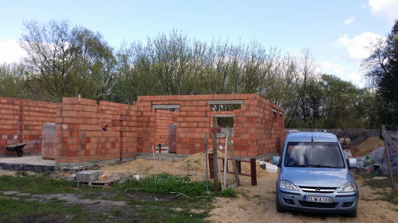 Wczesny etap budowy domu z czerwonej cegły, widoczne ściany w stanie surowym, zaparkowany niebieski samochód dostawczy na tle placu budowy.