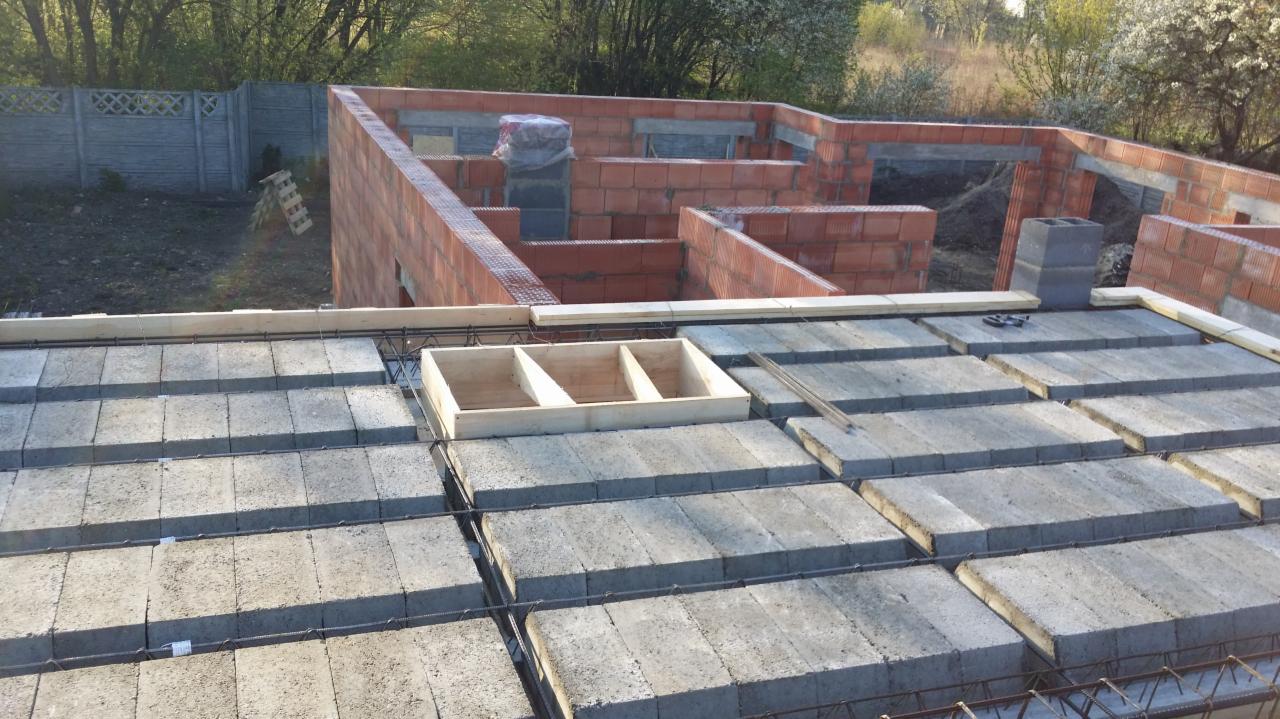 Wznoszenie ścian z czerwonej cegły na budowie domu jednorodzinnego, widoczne zbrojenie i betonowe pustaki stropowe, drewniana szalunkowa forma na stropie, w tle ogrodzenie z prefabrykatów betonowych.