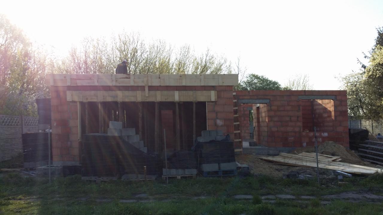 Wznoszenie ścian parterowego budynku z czerwonej cegły, widoczne drewniane szalunki i zbrojenia, pracownik na dachu, palety z bloczkami budowlanymi na pierwszym planie.