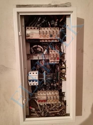 Otwarta, biała skrzynka rozdzielcza z widocznymi bezpiecznikami, przewodami i elementami montażowymi na szynach DIN; surowa ściana w tle.