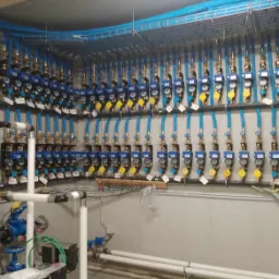 Rząd liczników wody z niebieskimi rurami i żółtymi zaworami zamontowanych na ścianie, z widocznymi elementami instalacji hydraulicznej w pomieszczeniu technicznym.