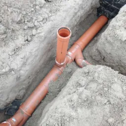 Pomarańczowe rury kanalizacyjne ułożone w wykopie ziemnym, widoczne złącza i pionowa rura wystająca z poziomej instalacji.