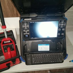 Hydra Usługi hydrauliczne - Kamera inspekcyjna z ekranem i klawiaturą, używana do diagnostyki rur. Czerwony detektor w otwartym futerale obok. Sprzęt na blacie.