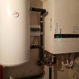 Hydra Usługi hydrauliczne - Biały bojler i piec gazowy zamontowane na ścianie, z widocznymi rurami i zaworami. Instalacja w pomieszczeniu o jasnych ścianach.