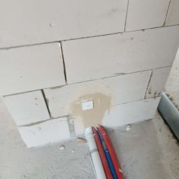 Hydra Usługi hydrauliczne - Instalacja wodna: rury (biała, czerwone, niebieska) przymocowane do ściany z betonu komórkowego, widoczna puszka elektryczna. Etap prac instalacyjnych.