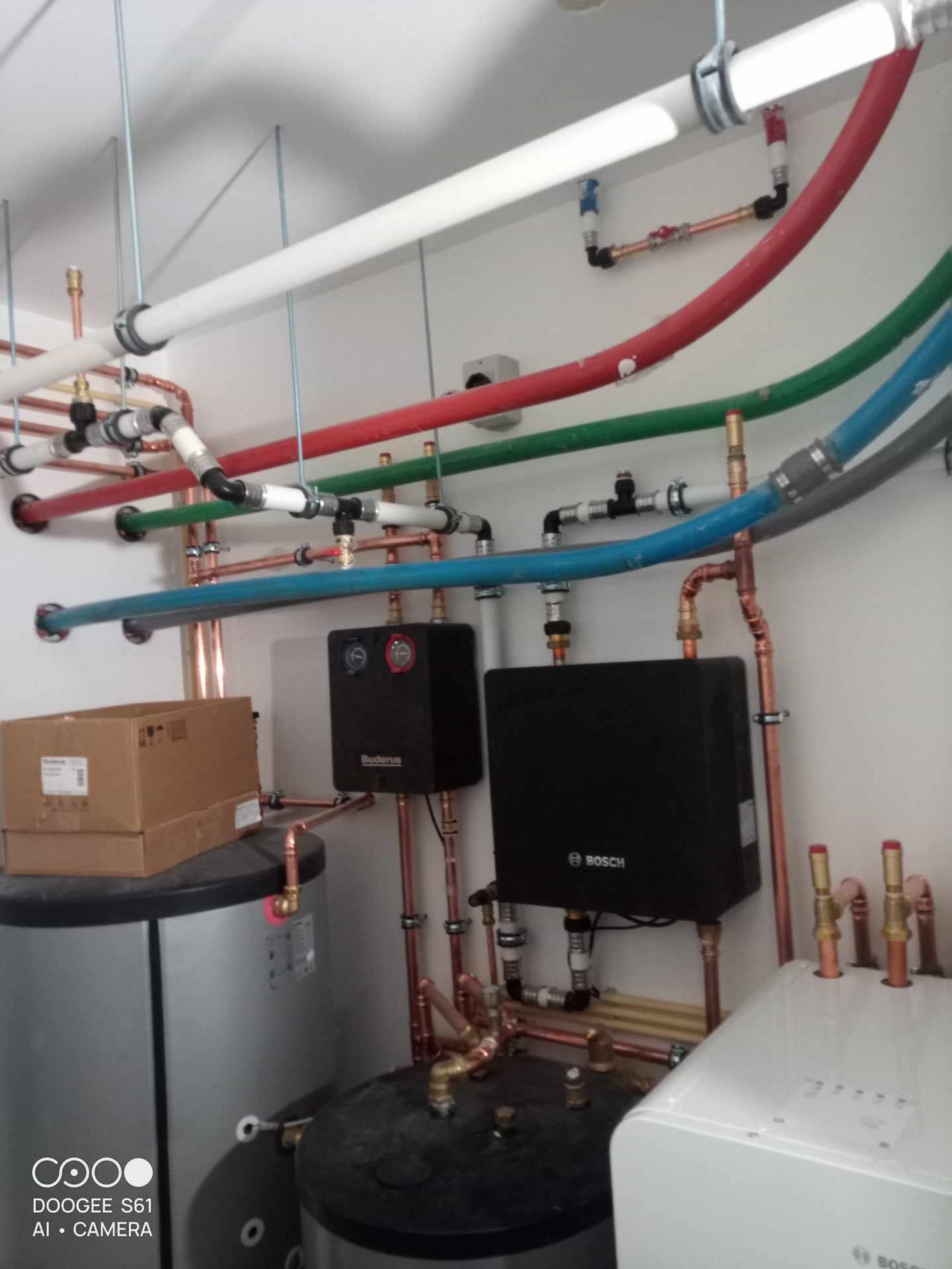 Złożona instalacja centralnego ogrzewania z kotłami Buderus i Bosch, rurami miedzianymi i plastikowymi w kolorach białym, czerwonym, zielonym i niebieskim, widok z dołu na instalację pod sufitem...