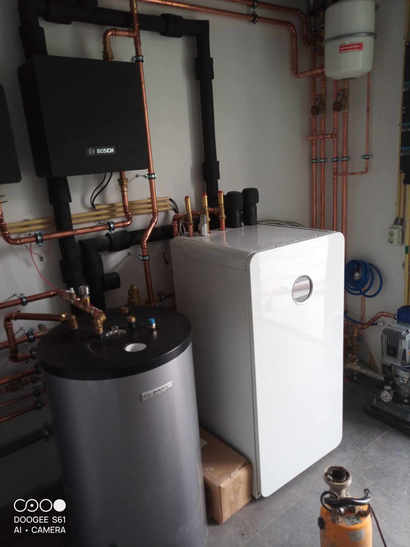 Nowoczesna kotłownia z dwoma zbiornikami, białym i szarym, połączonymi miedzianymi rurami z czarną izolacją, na ścianie widoczny moduł Bosch i naczynie przeponowe, na podłodze narzędzie hydrauliczne.