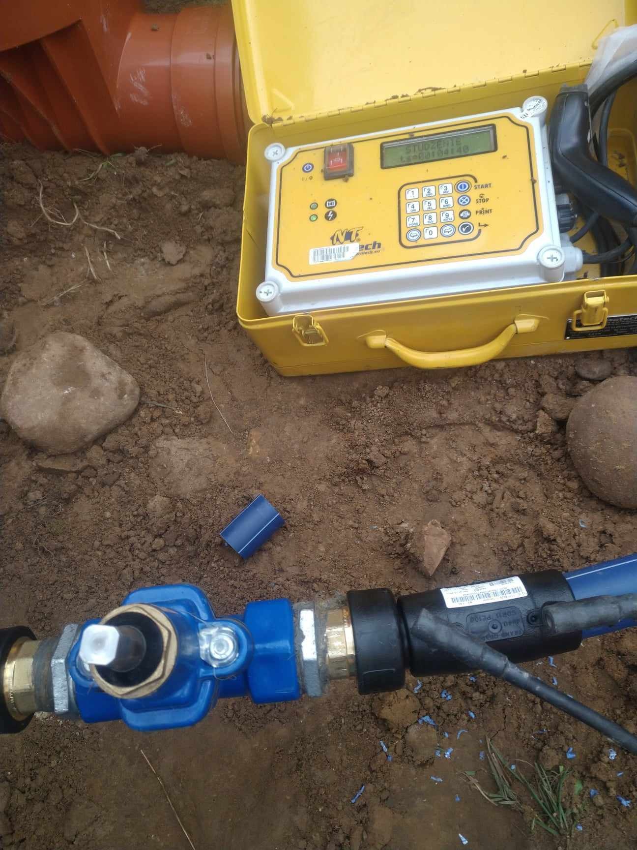 Niebieska rura wodociągowa z zaworem, podłączona do czarnego przewodu pomiarowego, leżąca w wykopie obok żółtej walizki z urządzeniem pomiarowym na ziemi.