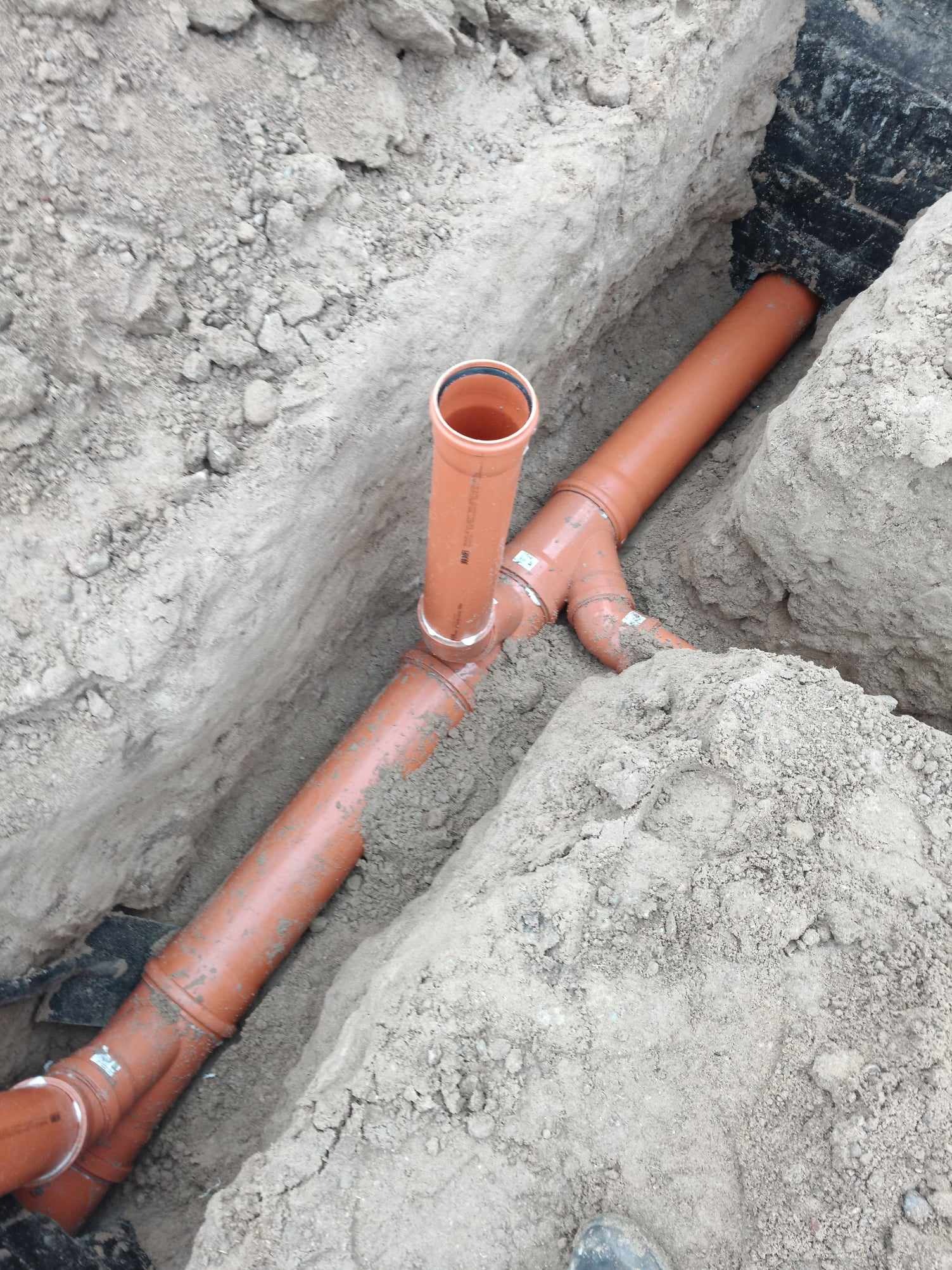 Pomarańczowe rury kanalizacyjne ułożone w wykopie ziemnym, widoczne złącza i pionowa rura wystająca z poziomej instalacji.