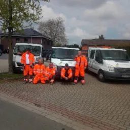 Zespół brukarzy w pomarańczowych kombinezonach pozuje na kostce brukowej przed trzema białymi samochodami dostawczymi z logo firmy ogrodniczej.
