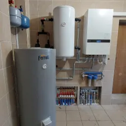 Kompletna instalacja grzewcza w kotłowni z szarym zasobnikiem Ferroli, białym bojlerem, białym piecem gazowym oraz rozdzielaczami ogrzewania podłogowego i filtrami wody.